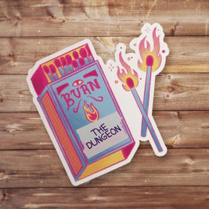 May include: A sticker set featuring a matchbox and two burning matches. The matchbox is blue with the word "BURN" and a flame graphic. The text "THE DUNGEON" is below. The matches have pink and yellow flames.