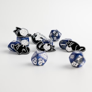 May include: A collection of blue and white polyhedral dice with black cat-shaped enamel pins. The pins feature a cat design with a white twenty-sided die. The dice are scattered on a white surface.