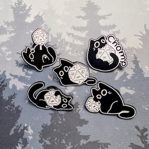 May include: Set of five black and white enamel pins featuring cartoon cats interacting with a twenty-sided die. One pin has the word "chomp" in white. The dice are white with black numbers. The cats have large eyes and are in various poses.