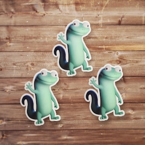 May include: Three cartoon lizard stickers in shades of green with white outlines are arranged on a wooden surface. Each lizard is waving with a friendly expression. The stickers have a glossy finish and are likely for decoration.