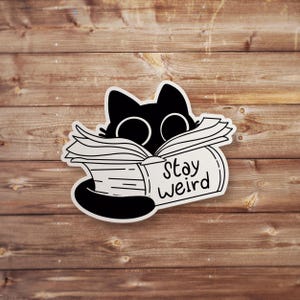 May include: A black and white sticker featuring a cartoon cat peeking over a book. The book has the words "Stay Weird" printed on it. The sticker is placed on a wooden background.