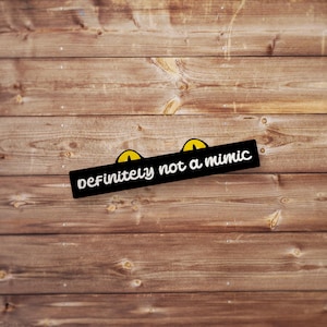 May include: Black and white sticker with yellow eyes and the text "definitely not a mimic" on a wood background.