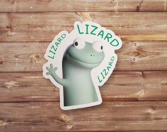May include: A sticker featuring a cartoon lizard in shades of green and white. The lizard is waving and the word "Lizard" is written around the image in green. The sticker is set against a wooden background.