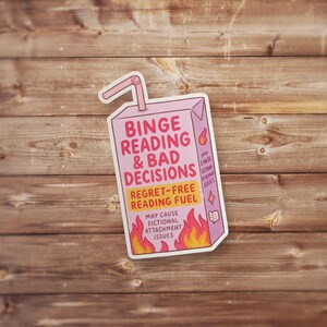May include: A sticker in the shape of a pink juice box with the text "Binge Reading & Bad Decisions" and "Regret-Free Reading Fuel." The sticker has a straw and flames at the bottom, with additional text about fictional attachment issues.