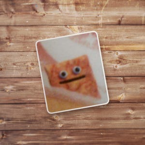 May include: A close-up of a sticker with a cartoon face on a wooden background. The face is made of a brown, textured material with two large, blue eyes and a black mouth.