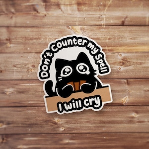 May include: A sticker featuring a black cartoon cat with large, teary eyes. The cat is holding a brown sign that reads "I will cry." Above the cat, the text "Don't counter my spell" is written in a circular pattern.
