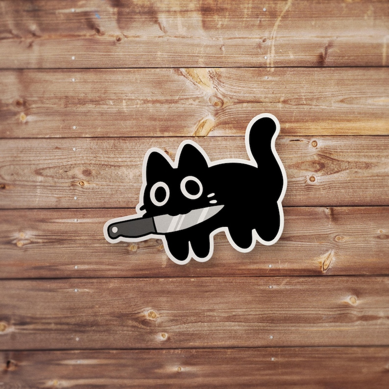 Pu&ograve; includere: Un adesivo di gatto nero con un coltello in bocca. Il gatto ha occhi grandi e rotondi e un design semplice e cartonato. L'adesivo &egrave; su uno sfondo di legno.