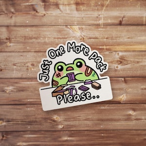 May include: A sticker featuring a cartoon frog with a green body, pink cheeks, and large black eyes. The frog is peeking over a white sign that reads "Just One More Pack Please.." in black text. The sticker is on a wooden surface.