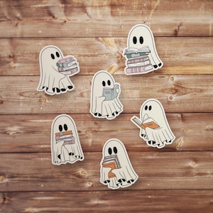 May include: Six white ghost stickers with black eyes and mouths, each holding a stack of books or a single book. The ghosts are on a wood background.