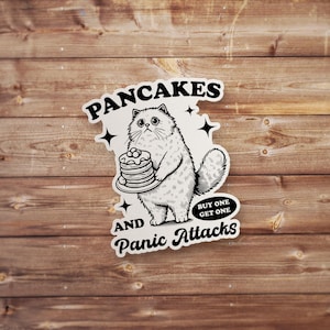 Pode incluir: Adesivo preto e branco com um gato de desenho animado segurando um prato de panquecas. O adesivo diz "Pancakes and Panic Attacks" com uma etiqueta "Buy One Get One".