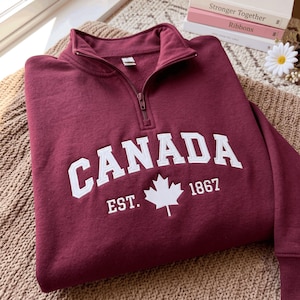 May include: A maroon quarter-zip sweatshirt featuring the white embroidered text CANADA EST. 1867 and a central maple leaf graphic. The garment has a high collar with a matching maroon zip pull and ribbed cuffs on the long sleeves.