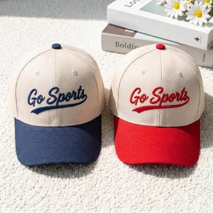 Può includere: Due cappellini da baseball con corona beige e testo ricamato che recita Go Sports. Un cappellino presenta visiera e scritte blu navy, mentre l'altro ha visiera e scritte rosse. Entrambi i cappellini sono dotati di un bottone superiore.