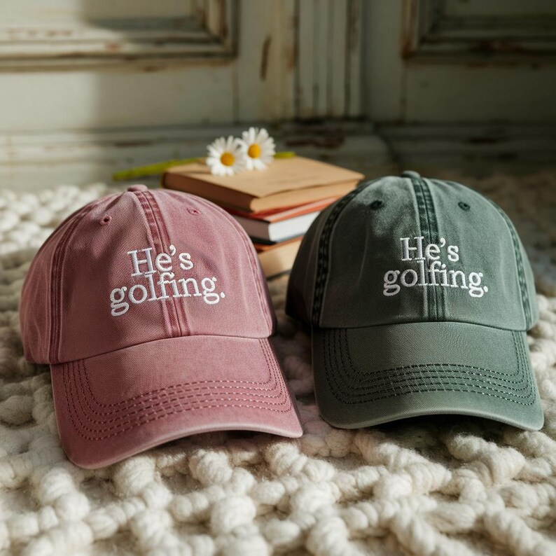 May include: Two baseball caps, one dusty rose and one olive green, are displayed. Both caps have the embroidered text "He's golfing." in white. The caps are sitting on a textured, off-white surface.