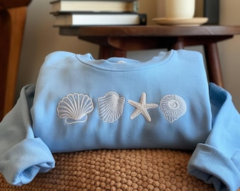Könnte beinhalten: Hellblaues Sweatshirt mit weißen, gestickten Muschel- und Seesternmotiven. Das Sweatshirt liegt auf einer braunen Oberfläche, im Hintergrund Bücher und eine Kaffeetasse. Das Design hat ein Küsten- oder Strandthema.