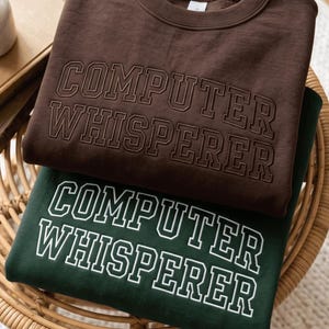 Puede incluir: Una sudadera de cuello redondo de color marrón oscuro con el texto COMPUTER WHISPERER bordado en hilo marrón oscuro a juego, sobre una sudadera de cuello redondo verde oscuro con el texto COMPUTER WHISPERER impreso en letras blancas.