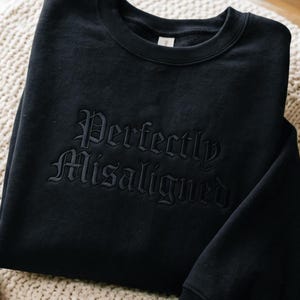 May include: A black crewneck sweatshirt featuring the words Perfectly Misaligned embroidered in a matching black thread across the chest. The text is rendered in a gothic-style font, creating a subtle, tonal appearance against the fabric.