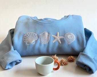Könnte beinhalten: Hellblaues Sweatshirt mit weißen Stickereien von Muscheln und Seesternen. Eine weiß-orange Tasse und kleine dekorative Elemente befinden sich vor dem Sweatshirt. Das Sweatshirt hat lange Ärmel.