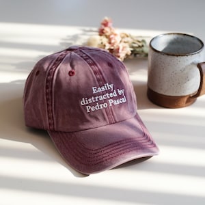 May include: A maroon baseball cap with the embroidered text "Easily distracted by Pedro Pascal" in white. The cap is next to a speckled mug with a brown base and handle, and a small bouquet of dried flowers.