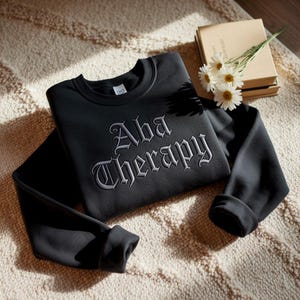 May include: Black crew neck sweatshirt with the embroidered text "Aba Therapy" in a gothic font. The sweatshirt is folded on a beige rug, with a stack of books and daisies in the background.