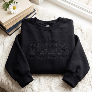 Könnte beinhalten: Ein schwarzer Rundhals-Pullover mit den Buchstaben BCBA in einer großen, fetten Schriftart im College-Stil, die auf die Brust gestickt ist. Unter den Buchstaben ist der Text Board Certified Behavior Analyst in einer kleineren Schreibschrift gestickt.