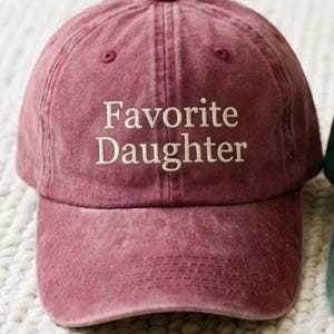 May include: A dusty rose baseball cap with the embroidered text "Favorite Daughter" in white. The cap has a curved brim and a fabric strap closure. The hat is made of a soft, textured material.