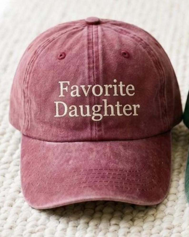 May include: A dusty rose baseball cap with the embroidered text "Favorite Daughter" in white. The cap has a curved brim and a fabric strap closure. The hat is made of a soft, textured material.
