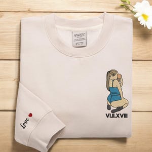 Może przedstawiać: Beżowa bluza z nadrukiem kobiety robiącej sobie selfie. Pod nadrukiem wyszyty jest tekst "VIII.II.XVIII". Na lewym rękawie wyszyty jest napis "Love" z czerwonym sercem.