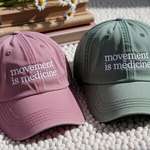 May include: Two baseball caps, one dusty rose and one sage green, are displayed. Each cap has the embroidered text "movement is medicine" in white. The caps are made of a soft fabric and have a curved brim, suitable for casual wear.