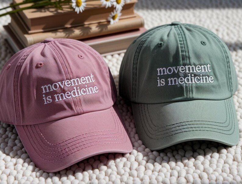 May include: Two baseball caps, one dusty rose and one sage green, are displayed. Each cap has the embroidered text "movement is medicine" in white. The caps are made of a soft fabric and have a curved brim, suitable for casual wear.