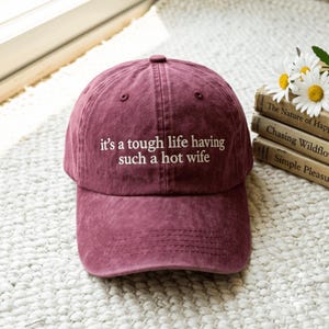 Puede incluir: Una gorra de béisbol color granate con textura de tela desgastada que presenta el texto bordado en blanco "it's a tough life having such a hot wife" centrado en el panel frontal, con visera curva y un botón en la parte superior.