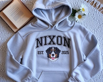 May include: A light gray hoodie with a front pocket and hood. The word "NIXON" is printed across the chest in black block letters, with "EST. 2025" below. A dog's face is centered below the text. A book and flowers are in the background.