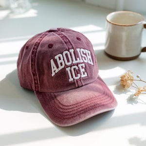 May include: A burgundy baseball cap with the text "ABOLISH ICE" in white block letters. The cap has a curved brim and a distressed, vintage look. The hat is made of a soft, worn material.