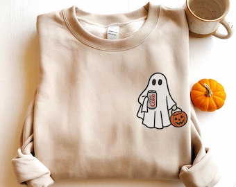 Op de afbeelding: Een beige sweater met ronde hals en een afbeelding van een witte geest die een blikje Diet Coke en een oranje pompoenemmer vasthoudt. Het ontwerp bevindt zich op de linkerborst van het kledingstuk, dat is voorzien van lange mouwen.