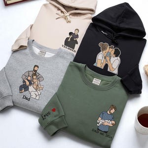 May include: A selection of sweatshirts and a hoodie in beige, grey, black, and olive green. Each item has an embroidered family portrait design. The designs show parents with children, and the word "Dad" is visible on one.