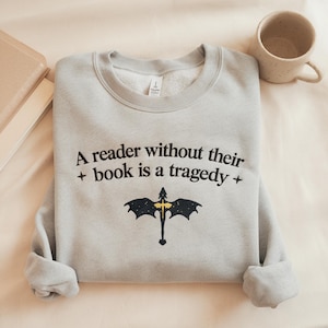 May include: Light gray sweatshirt with the text "A reader without their book is a tragedy" and a black and gold graphic. A speckled mug and a book are in the background.