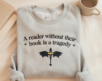 Può includere: Felpa grigio chiaro con la scritta "A reader without their book is a tragedy" e una grafica nera e oro. Una tazza maculata e un libro sono visibili sullo sfondo.
