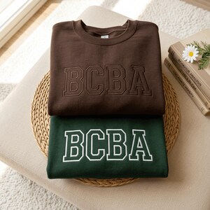 May include: Two crewneck sweatshirts stacked on top of each other. The top sweatshirt is dark brown with the letters BCBA embroidered in a matching dark brown thread. The bottom sweatshirt is dark green with the letters BCBA embroidered in white.