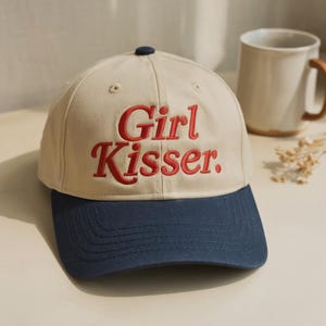 May include: A beige baseball cap with a dark blue brim and a dark blue button on top. The front of the cap features the text Girl Kisser. embroidered in a bold red serif font. The cap has a structured crown with stitched eyelets for ventilation.