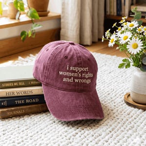 以下が含まれることがあります： フロントパネルに「i support women's rights and wrongs」という白い刺繍が施された、色あせた風合いのマルーンカラーの野球帽。カーブしたつばと、上部に通気用のアイレットを備えた構造的なクラウンが特徴です。