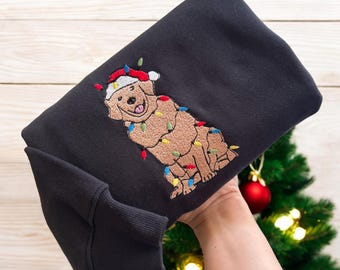 May include: Black sweatshirt featuring an embroidered golden retriever wearing a Santa hat and Christmas lights. The dog is smiling with its tongue out. A red Christmas ornament and a small Christmas tree are in the background.