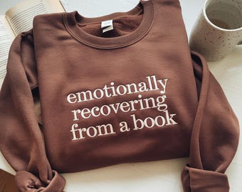 May include: A brown sweatshirt with the embroidered text "emotionally recovering from a book" in white. The sweatshirt is folded on a white surface, with a book and a mug in the background.