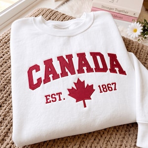 May include: A white crewneck sweatshirt featuring red embroidered text that reads CANADA in an arched font. Below the text is a red embroidered maple leaf flanked by the text EST. on the left and 1867 on the right, all in a matching red font.