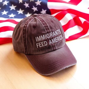 May include: A maroon baseball cap with the text "IMMIGRANTS FEED AMERICA" in white. The cap is set against the backdrop of an American flag, with its red and white stripes and stars visible.