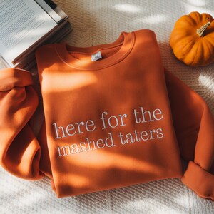 May include: Orange sweatshirt with the phrase "here for the mashed taters" embroidered in white. A small pumpkin and a book are in the background. The sweatshirt is folded on a white surface.