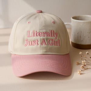 May include: A baseball cap with a cream-colored crown and a pink bill. The cap features the embroidered text "Literally Just A Girl" in pink. The cap has a pink button on top.