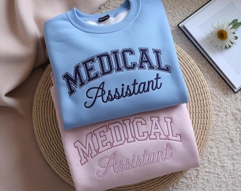 May include: Two crew neck sweatshirts, one pale blue and one pink, are stacked. Each has the embroidered text "MEDICAL Assistant" in a collegiate font. A book and daisy are in the background. The sweatshirts are on a woven mat.