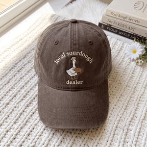 May include: A dark brown baseball cap featuring an embroidered graphic of a white goose wearing a chef hat and holding a loaf of bread. The text embroidered in a cream colour above and below the goose reads local sourdough dealer.
