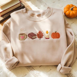 Puede incluir: Sudadera beige con diseños bordados con temática otoñal. Presenta una taza de café con un corazón, un libro, una hoja, una vela y una calabaza. Una pequeña calabaza naranja en el fondo.