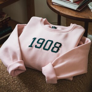 May include: A pink sweatshirt with a dark green embroidered number "1908" on the front.