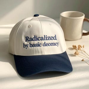 May include: A white and navy blue baseball cap with the embroidered text "Radicalized by basic decency." The cap has a curved brim and a small navy blue button on top. A beige mug with a brown handle is in the background.